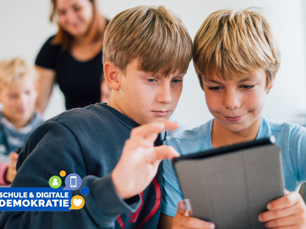 KI generiert: Das Bild zeigt zwei Jungen, die gemeinsam auf ein Tablet schauen, während sie in einer Gruppe mit anderen Kindern und einer erwachsenen Person im Hintergrund sind. Der Hauptinhalt des Bildes ist die Interaktion der Kinder mit digitalen Medien, was durch den Text "Schule & digitale Demokratie" betont wird.