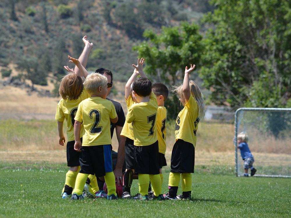 KI generiert: Das Bild zeigt eine Gruppe von Kindern in gelben Sporttrikots, die auf einem Feld stehen und sich mit hochgereckten Armen freuen. Im Hintergrund sind Bäume, ein Haus und ein Tor zu erkennen.