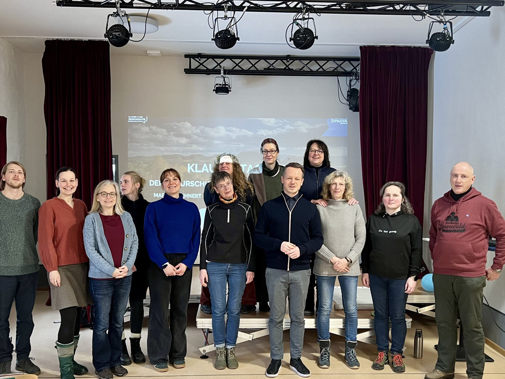 KI generiert: Das Bild zeigt eine Gruppe von Personen, die vor einem Projektorbildschirm mit der Aufschrift "KLAUSURTAG" posieren. Der Hauptinhalt des Bildes ist eine Gruppenaufnahme, die vermutlich im Rahmen einer Klausurtagung oder eines ähnlichen Anlasses entstanden ist.