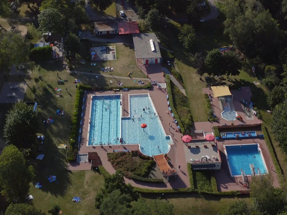 KI generiert: Das Bild zeigt eine Luftaufnahme eines Freizeitbades mit mehreren Schwimmbecken, umgeben von Grünflächen und Gebäuden. Der Hauptinhalt des Bildes ist das Schwimmbad mit seinen verschiedenen Becken und den umliegenden Freizeiteinrichtungen.
