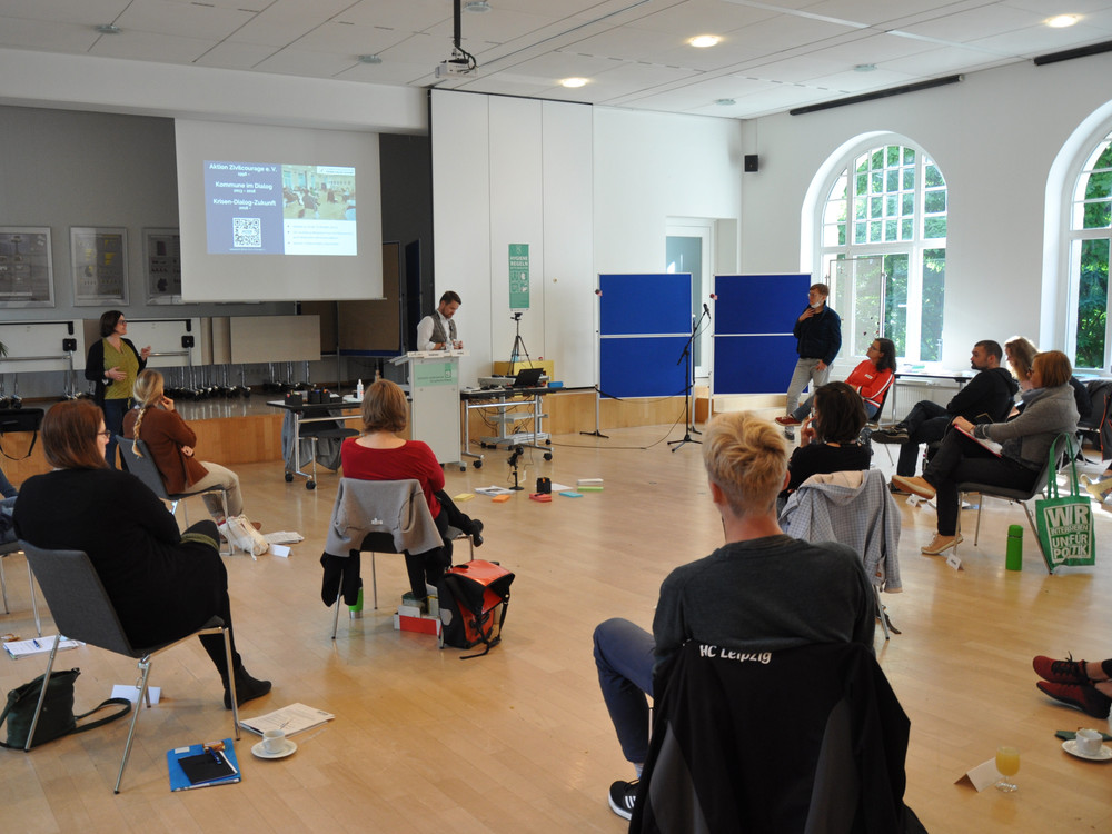 KI generiert: Das Bild zeigt eine Gruppe von Personen in einem hellen Raum, die an einer Präsentation oder einem Workshop teilnehmen. Im Vordergrund sitzen die Zuhörer, während im Hintergrund zwei Personen vor einem Podium und einer Leinwand stehen und sprechen.