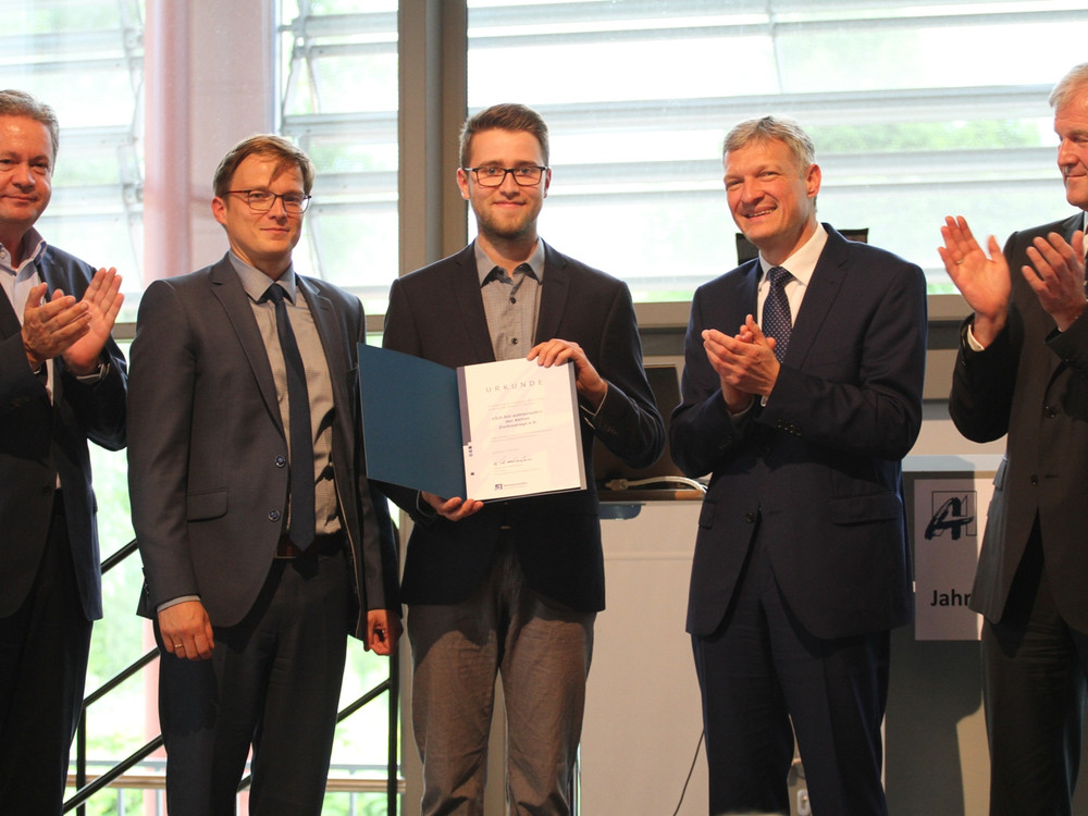 KI generiert: Das Bild zeigt eine Gruppe von fünf Männern in formeller Kleidung, wobei einer von ihnen eine Urkunde oder ein Buch in den Händen hält. Die anderen vier applaudieren, was darauf hindeutet, dass eine Auszeichnung oder Anerkennung stattfindet.