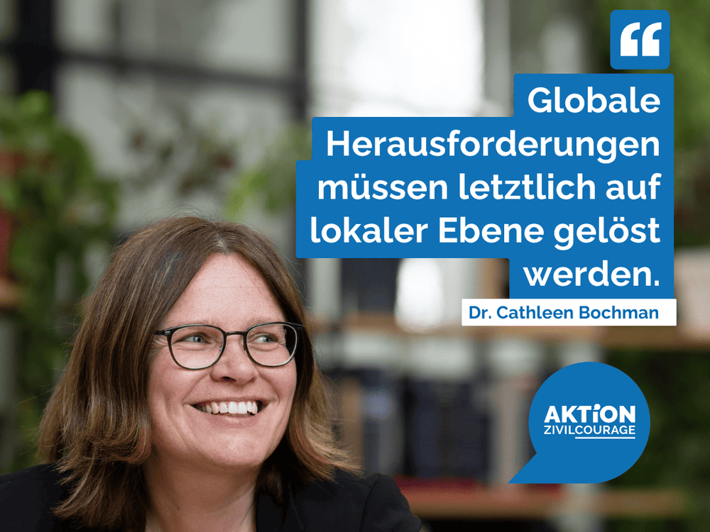 KI generiert: Das Bild zeigt eine lächelnde Frau mit Brille, die vor einem Bücherregal sitzt. Im Vordergrund ist ein blaues Sprechblasen-Symbol mit der Aufschrift "AKTION ZIVILCOURAGE" zu sehen.