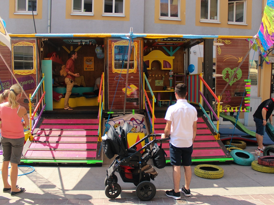 KI generiert: Das Bild zeigt eine bunte, mobile Spiel- und Kletteranlage für Kinder, die auf einem Platz vor einem Gebäude aufgestellt ist. Mehrere Erwachsene und Kinder sind in der Nähe, einige beobachten die Anlage oder bereiten sich darauf vor, sie zu nutzen.