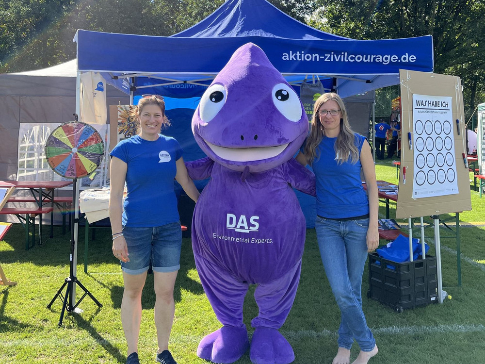 KI generiert: Das Bild zeigt zwei Personen in blauen T-Shirts, die neben einem großen lila Maskottchen in Form eines Tropfens stehen, vor einem Stand mit der Aufschrift "aktion-zivilcourage.de". Im Hintergrund sind ein Glücksrad und ein Plakat mit der Aufschrift "WAS HABE ICH?" zu sehen.