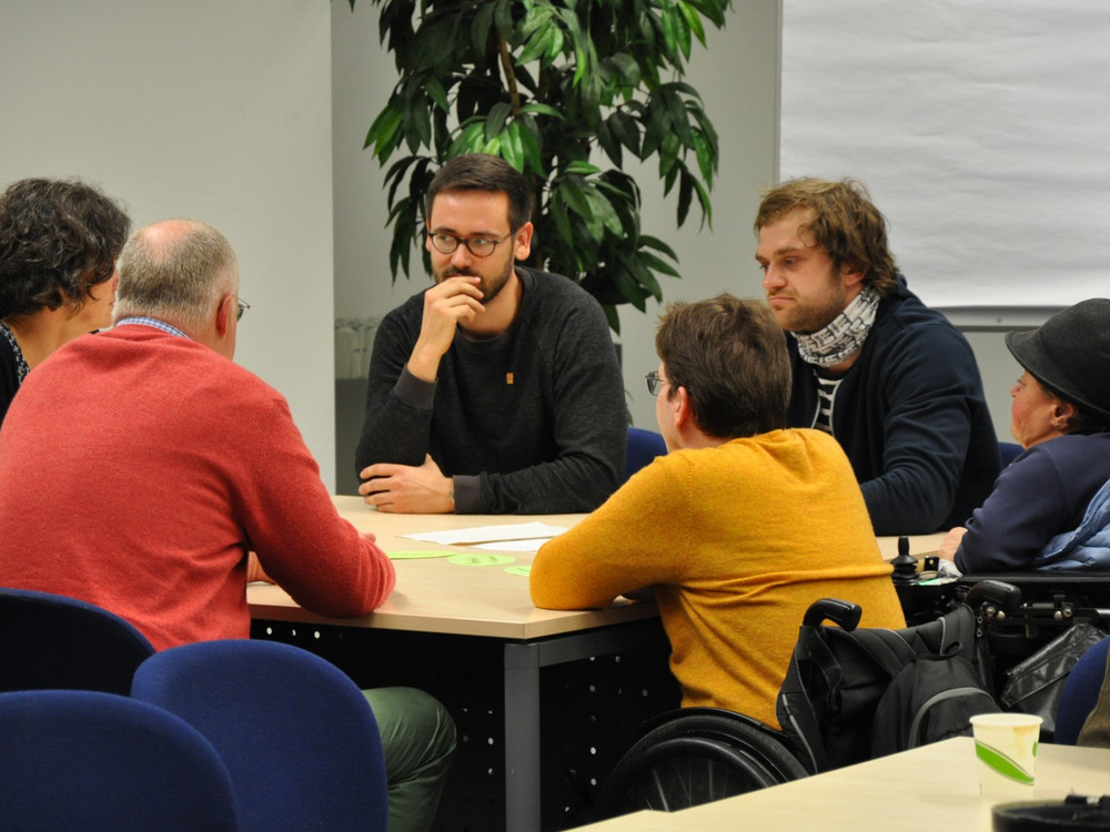 KI generiert: Das Bild zeigt eine Gruppe von Menschen, die an einem Tisch sitzen und sich in einer Besprechung oder Diskussion befinden. Der Hauptinhalt des Bildes ist eine Meeting-Situation in einem Büro oder Seminarraum.