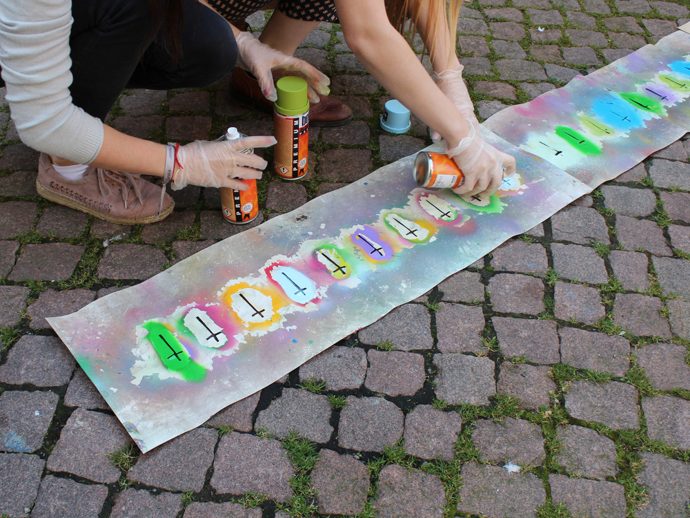 KI generiert: Das Bild zeigt mehrere Personen, die auf einem cobbelsteinbelagten Boden mit Sprühdosen bunte Graffiti auf eine lange Papierbahn sprühen. Der Hauptinhalt des Bildes ist das gemeinsame kreativ-künstlerische Gestalten von Graffiti.