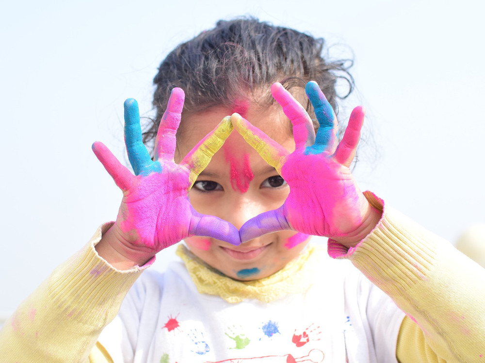 KI generiert: Das Bild zeigt ein Kind mit bunt bemalten Händen, das durch einen Rahmen aus seinen Händen schaut und dabei lächelt. Der Hauptinhalt des Bildes ist die kreative und spielerische Darstellung des Kindes mit den farbenfrohen Handabdrücken.
