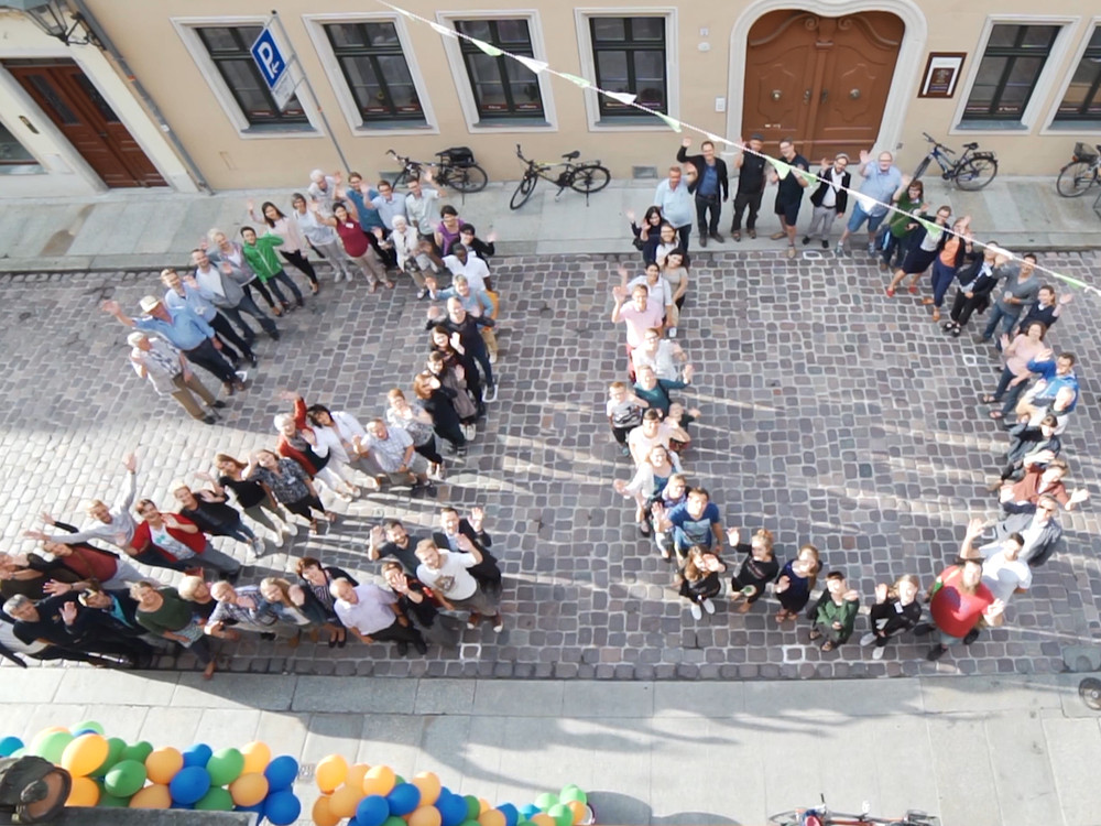 KI generiert: Das Bild zeigt eine große Gruppe von Menschen, die sich auf einem gepflasterten Platz zu einer Formation aufgestellt haben, um ein Wort oder eine Form zu bilden. Im Vordergrund sind bunte Ballons zu sehen, die an einer Absperrung befestigt sind.