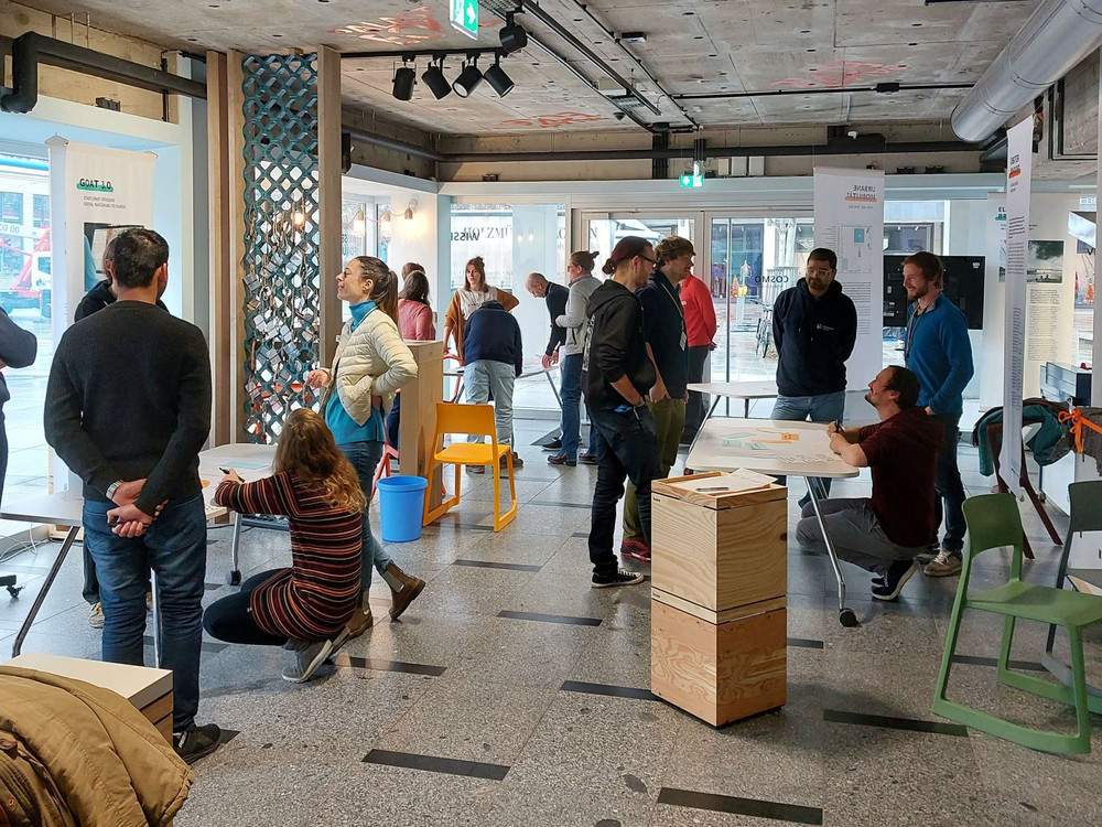 KI generiert: Das Bild zeigt eine Gruppe von Menschen in einem hellen, modernen Innenraum, die sich unterhalten und an Tischen stehen. Der Hauptinhalt des Bildes ist eine soziale Zusammenkunft oder ein Event in einem Gemeinschaftsraum oder Büro.