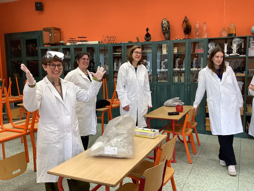 KI generiert: Das Bild zeigt fünf Personen in weißen Laborkitteln, die in einem Labor stehen und fröhlich in die Kamera winken. Im Hintergrund sind Laborgeräte, Regale mit Chemikalien und andere typische Laborausstattungen zu sehen.