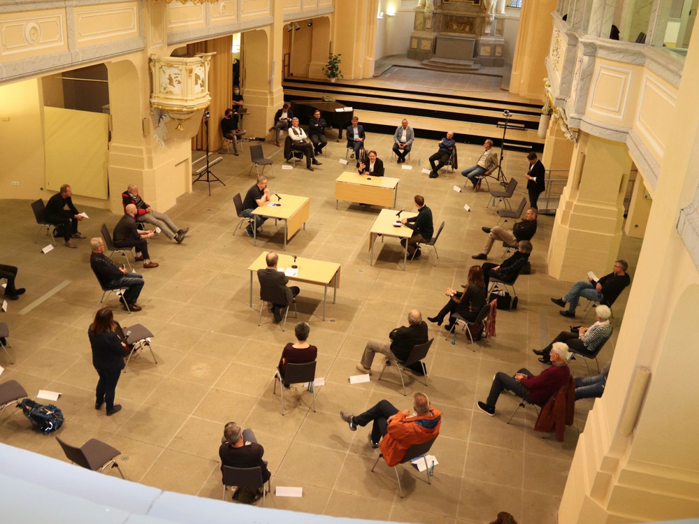 KI generiert: Das Bild zeigt eine Versammlung oder ein Meeting in einem großen, hellen Raum mit hohen Decken und Säulen. Im Mittelpunkt stehen mehrere Personen an Tischen mit Mikrofonen, während andere im Publikum sitzen und zuhören.