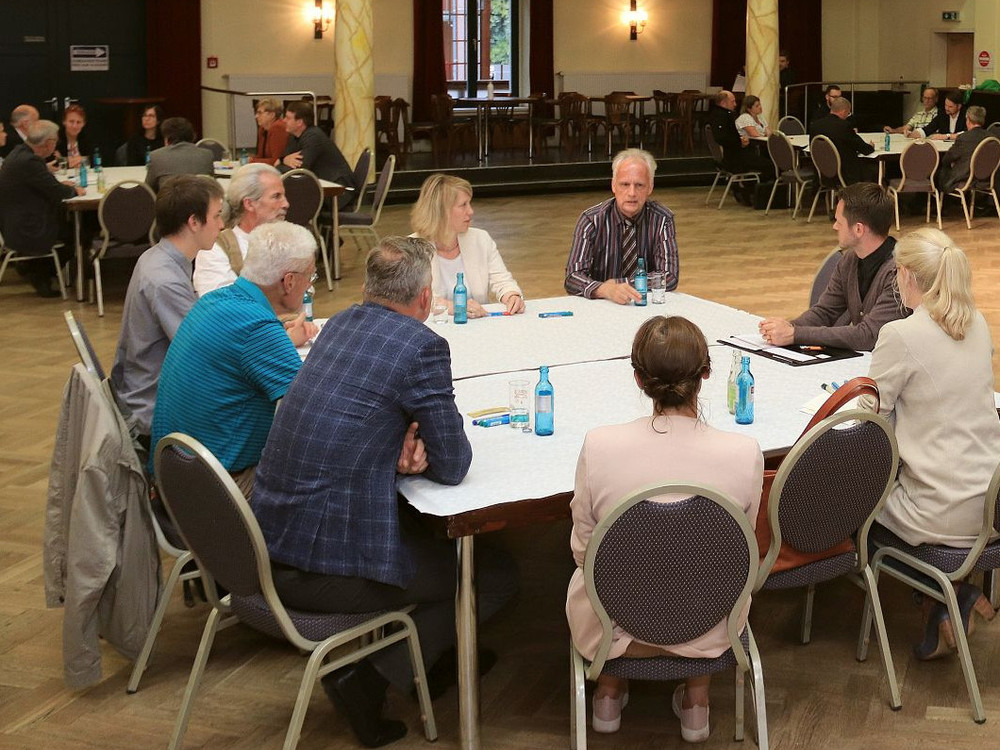 KI generiert: Das Bild zeigt eine Gruppe von Personen in einem hellen Raum, die einem Vortragenden zuhören, der vor einem Flipchart mit bunten Notizen steht. Der Hauptinhalt des Bildes ist eine Präsentation oder ein Workshop in einer informellen Lernumgebung.
