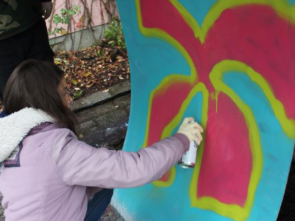 KI generiert: Das Bild zeigt eine Person, die auf einem großen, blauen Holzbrett ein Graffiti in Form einer roten Blume mit gelben Konturen sprüht. Die Szene spielt im Freien, möglicherweise in einem Park oder einer urbanen Umgebung.