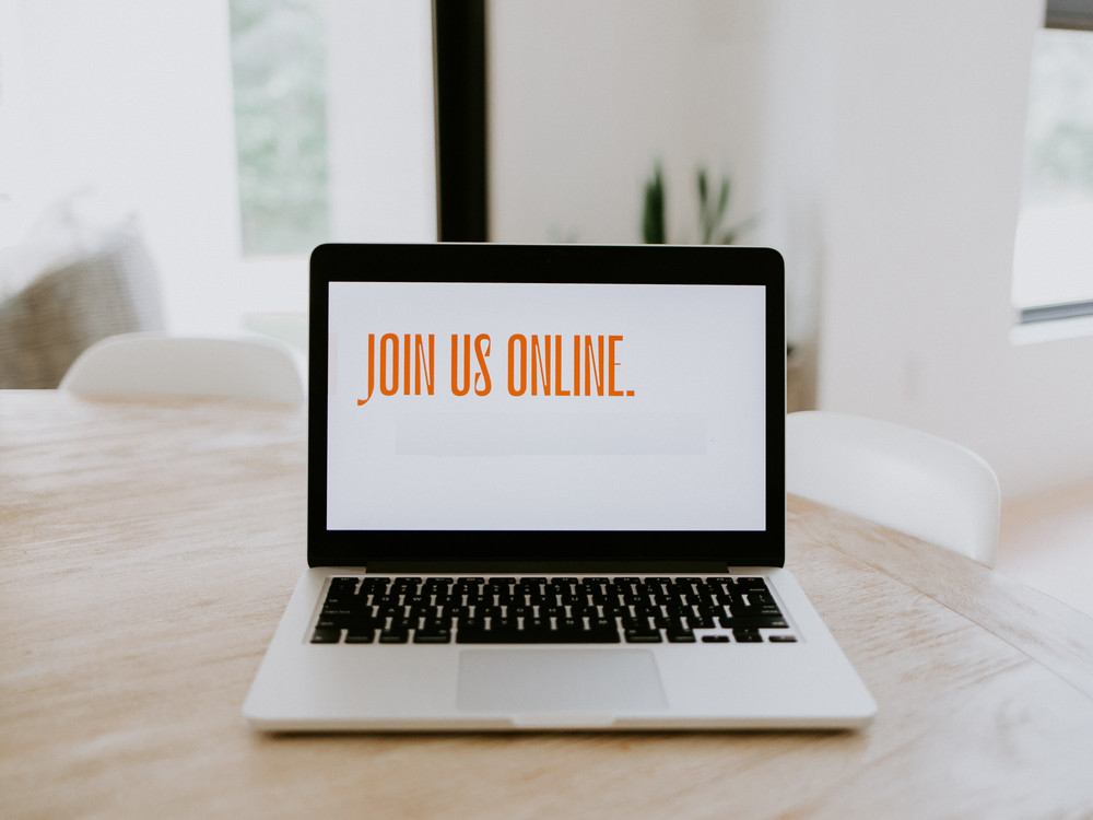 KI generiert: Das Bild zeigt einen Laptop auf einem Holztisch mit der Bildschirmanzeige "JOIN US ONLINE." Im Hintergrund sind ein Stuhl und ein Fenster zu sehen. Der Hauptinhalt des Bildes ist die Einladung, online teilzunehmen.