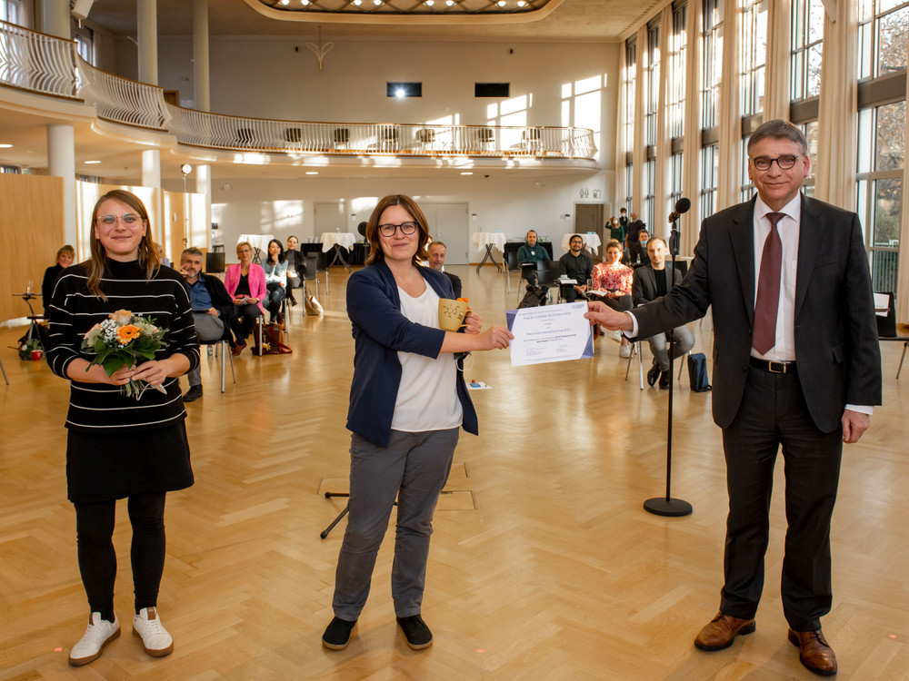 KI generiert: Das Bild zeigt eine offizielle Zeremonie in einem hellen, geräumigen Raum, bei der eine Frau von einem Mann in Anzug eine Urkunde und eine kleine Trophäe erhält, während eine weitere Frau mit Blumen im Arm daneben steht. Im Hintergrund sind mehrere Personen als Publikum anwesend.