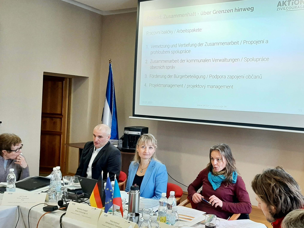 KI generiert: Das Bild zeigt eine formelle Besprechung oder Konferenz in einem gut beleuchteten Raum mit mehreren Teilnehmern, die an langen Tischen sitzen. Im Vordergrund steht eine Person, die eine Präsentation hält, während im Hintergrund eine Leinwand mit dem Titel "Projektpartnertreffen" zu sehen ist.