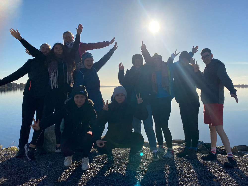 KI generiert: Das Bild zeigt eine Gruppe von Menschen, die draußen in der Natur stehen und fröhlich in die Kamera winken. Im Hintergrund ist ein See und ein klarer Himmel mit strahlendem Sonnenschein zu sehen.
