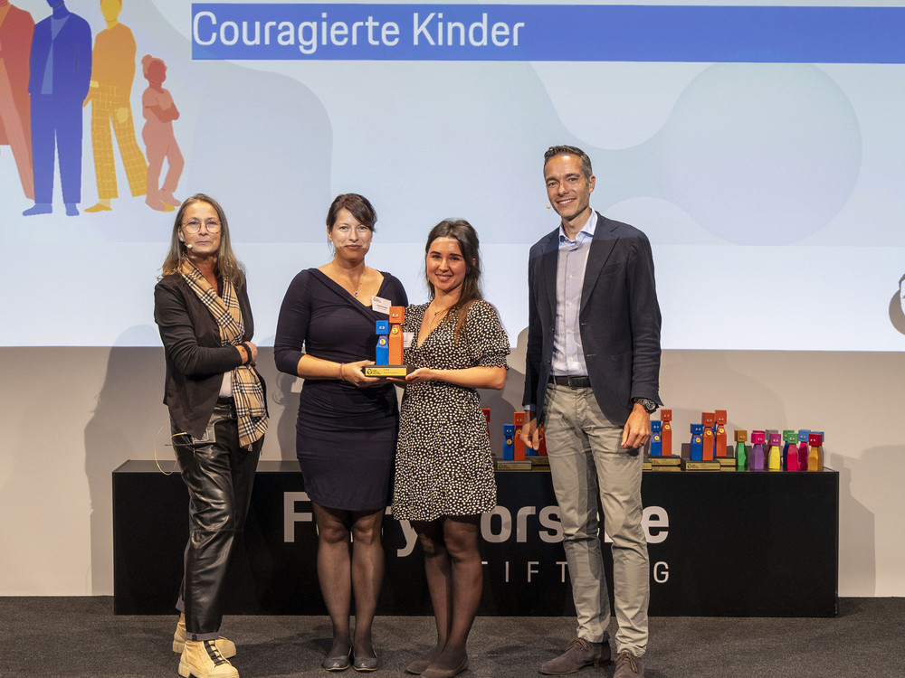 KI generiert: Das Bild zeigt eine große Gruppe von Menschen, die auf einer Bühne stehen und gemeinsam posieren. Viele von ihnen halten eine orangefarbene Urkunde oder einen Gegenstand in der Hand, und im Hintergrund ist ein Banner mit der Aufschrift "ZUSAMMENLEBEN" zu sehen.