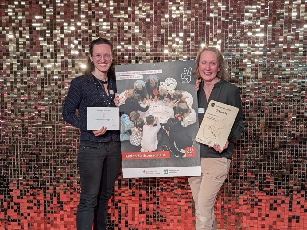 KI generiert: Das Bild zeigt zwei lächelnde Frauen, die jeweils eine Urkunde und ein Buch in den Händen halten, vor einer glänzenden, mosaikartigen Wand. Der Hauptinhalt des Bildes ist die Verleihung oder Anerkennung, die durch die Urkunden symbolisiert wird.