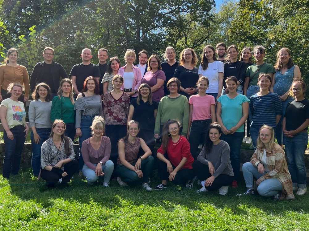 KI generiert: Das Bild zeigt eine Gruppe von fünf Personen, die an einem Tisch sitzen und sich in einer Diskussion oder einem Meeting befinden. Der Hauptinhalt des Bildes ist eine Besprechung oder ein Workshop in einem hellen Raum mit großen Fenstern und Blick auf einen Garten.