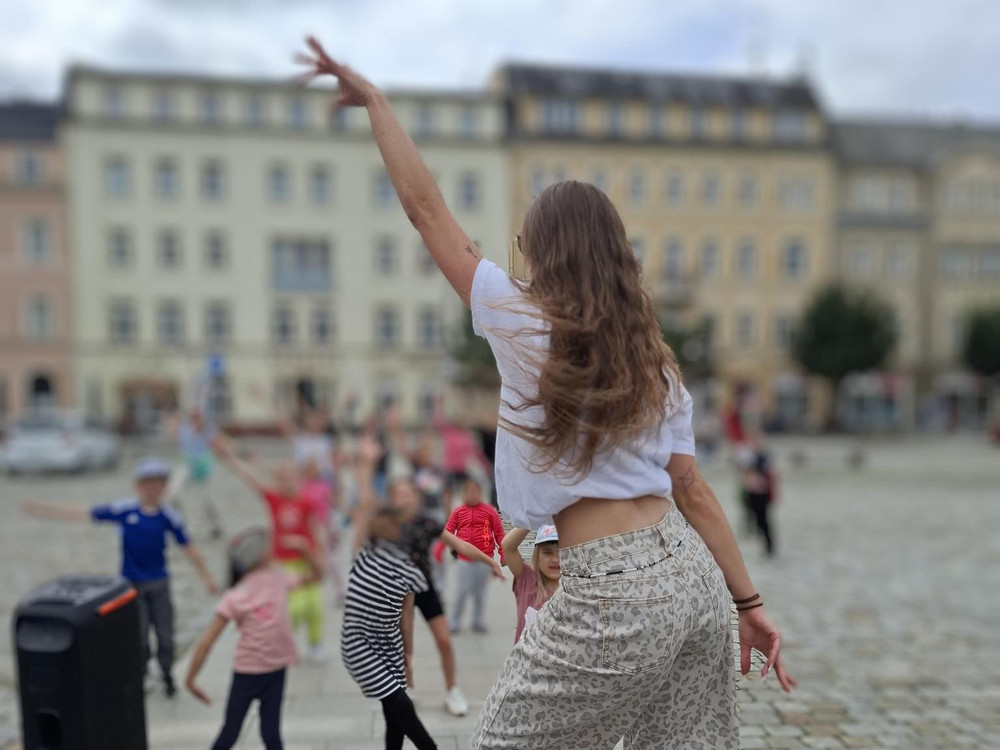 KI generiert: Das Bild zeigt eine Gruppe von Kindern und Jugendlichen, die draußen eine Tanz- oder Bewegungsaktivität durchführen, angeleitet von einer erwachsenen Person. Im Hintergrund sind Gebäude und Bäume zu sehen, was darauf hindeutet, dass die Aktivität in einem städtischen Umfeld stattfindet.