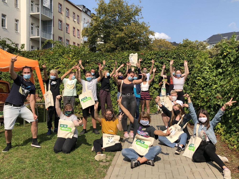 KI generiert: Das Bild zeigt eine Gruppe von Menschen, die draußen vor einer grünen Hecke stehen und sich freudig mit hochgestreckten Armen präsentieren, wobei viele von ihnen Gesichtsmasken tragen. Der Hauptinhalt des Bildes ist eine fröhliche Gruppenaktivität, möglicherweise im Rahmen einer gemeinnützigen oder ökologischen Veranstaltung.