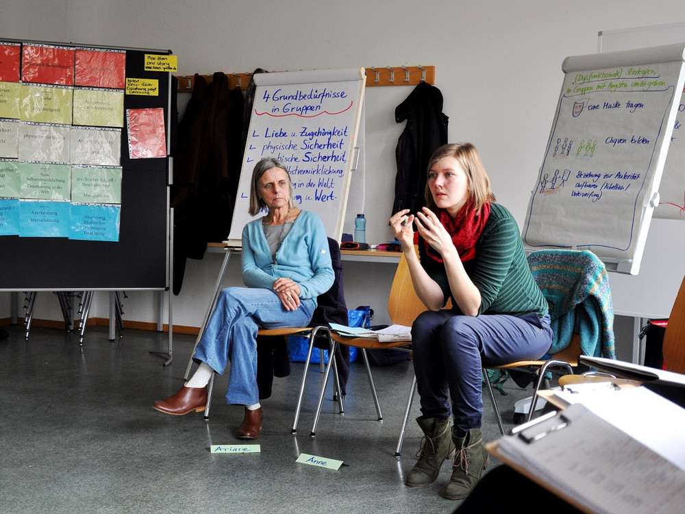 KI generiert: Das Bild zeigt zwei Frauen in einem Seminarraum, die auf Stühlen sitzen und an einer Gruppenarbeit oder Diskussion teilnehmen. Im Hintergrund sind Flipcharts mit Notizen und Plänen zu sehen, die auf eine organisierte Arbeits- oder Lernsituation hinweisen.