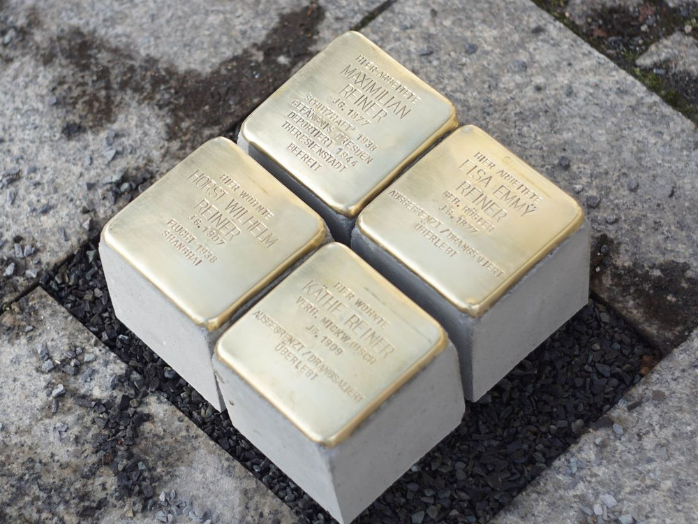 KI generiert: Das Bild zeigt eine Hand, die eine goldene Gedenktafel auf einem Stein berührt, umgeben von zwei weißen Rosen. Der Hauptinhalt des Bildes ist eine Hommage oder Erinnerung an eine Person oder ein Ereignis, wie durch die Gedenktafel und die Blumen symbolisiert wird.