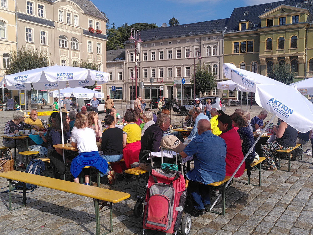 KI generiert: Das Bild zeigt eine belebte Außenbereichs-Szene mit vielen Menschen, die an Tischen unter großen, weißen Sonnenschirmen mit der Aufschrift "AKTION" sitzen. Im Hintergrund sind Gebäude und weitere Personen zu sehen, was auf eine öffentliche Veranstaltung oder ein Café in einer Stadt hinweist.