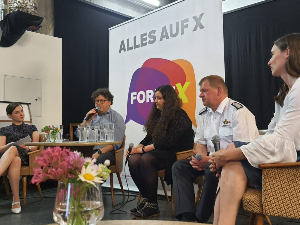 KI generiert: Das Bild zeigt eine Podiumsdiskussion mit fünf Personen, die auf der Bühne sitzen und Mikrofone verwenden. Im Hintergrund ist ein Banner mit der Aufschrift "FORUM X" zu sehen.