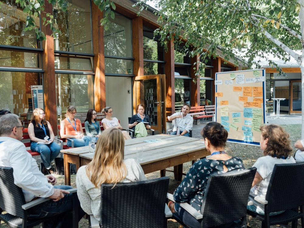 KI generiert: Das Bild zeigt eine Gruppe von Menschen, die im Freien an einem großen Holztisch sitzen und sich in einer Besprechung oder einem Workshop befinden. Im Hintergrund sind ein Gebäude mit großen Fenstern und ein Whiteboard mit Notizen zu sehen.