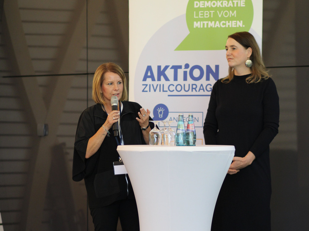 KI generiert: Das Bild zeigt eine Frau, die vor einem Publikum spricht und dabei gestikuliert. Der Hauptinhalt des Bildes ist eine Person, die eine Präsentation oder Rede hält.