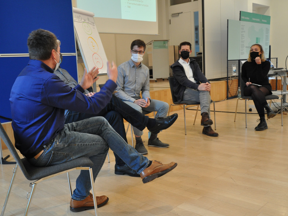 KI generiert: Das Bild zeigt eine Gruppe von Personen in einem Seminarraum, wobei eine Person in einem blauen Jackett steht und eine Präsentation hält, während die anderen Teilnehmer, die alle Masken tragen, zuhören. Der Hauptinhalt des Bildes ist eine interaktive Präsentation oder Diskussion in einem geschäftlichen oder akademischen Umfeld.