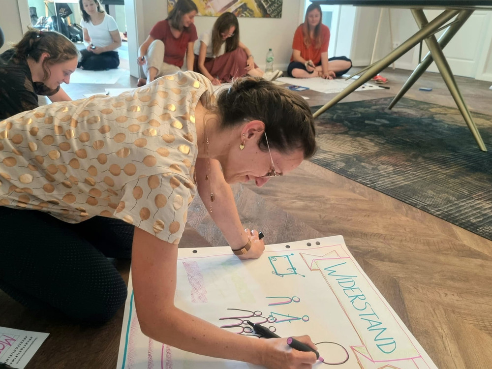 KI generiert: Das Bild zeigt eine Gruppe von Personen, die in einem hellen, modernen Raum mit großen Fenstern und Pflanzen sitzen und einer Präsentation folgen. Der Hauptinhalt ist eine Schulung oder ein Workshop, bei dem ein Mann vorne steht und etwas an einem Flipchart erklärt.