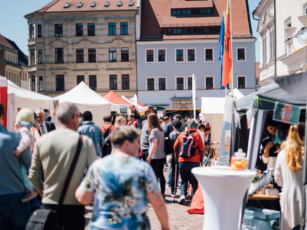 KI generiert: Das Bild zeigt eine belebte Straßenveranstaltung mit mehreren Ständen, darunter ein Stand mit der Aufschrift "Stück des Weges". Menschen schlendern durch die Gassen, betrachten die Angebote und interagieren miteinander.