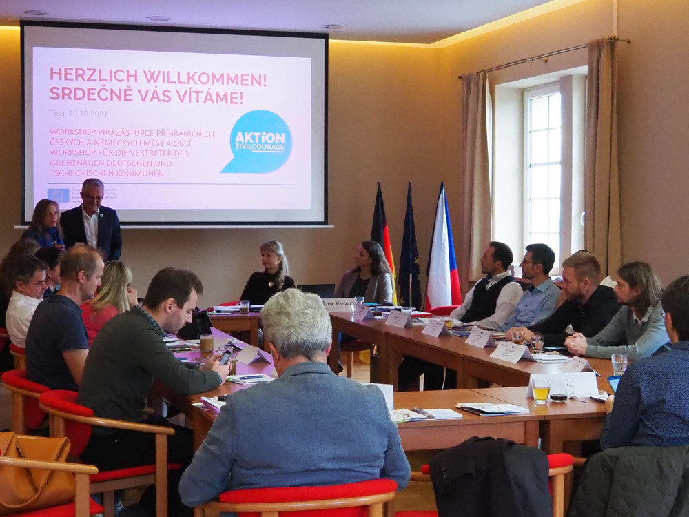 KI generiert: Das Bild zeigt eine formelle Sitzung oder Konferenz in einem Raum mit mehreren Personen, die an einem langen Tisch sitzen. Im Hintergrund sind zwei Flaggen, die deutsche und die tschechische, sowie eine Person, die vorne steht und möglicherweise eine Präsentation hält.