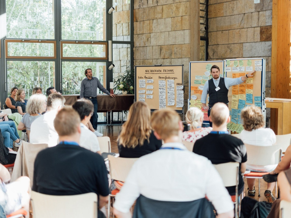 KI generiert: Das Bild zeigt eine Gruppe von Menschen in einem Seminarraum, wobei zwei Männer vorne stehen und eine Präsentation halten. Der Hauptinhalt des Bildes ist eine Schulung oder ein Workshop, bei dem die Teilnehmer aufmerksam zuhören und Notizen machen.