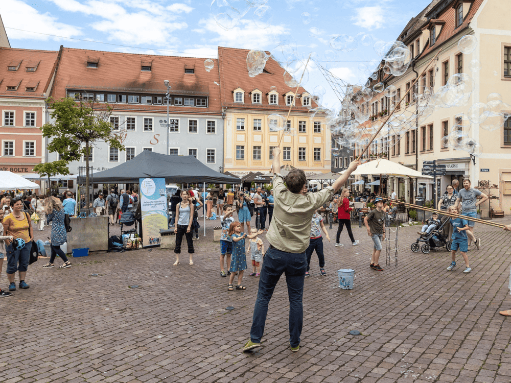 KI generiert: Das Bild zeigt eine belebte Platzszene mit Menschen, die sich an verschiedenen Aktivitäten beteiligen, darunter das Erzeugen großer Seifenblasen. Im Hintergrund sind bunte Gebäude und Marktstände zu sehen, die eine lebendige Atmosphäre schaffen.