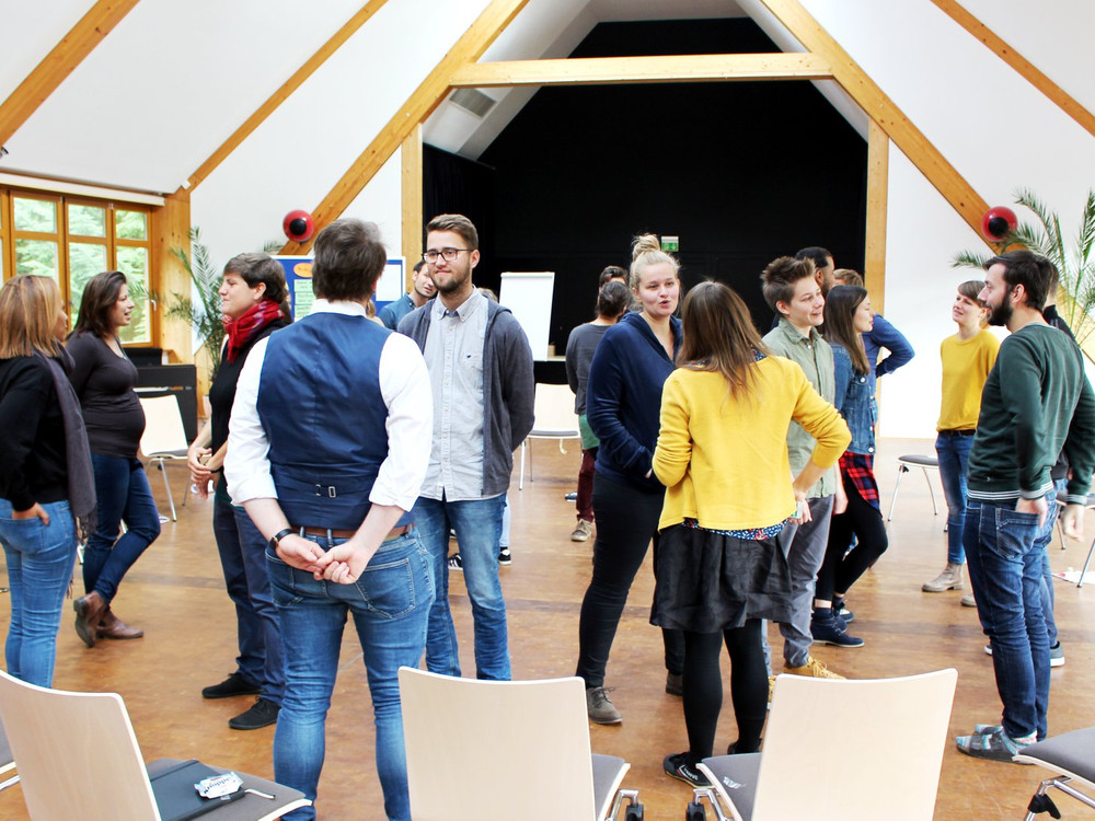 KI generiert: Das Bild zeigt eine Gruppe von Menschen, die in einem hellen, geräumigen Raum mit Holzvertäfelung und großen Fenstern stehen und sich unterhalten. Der Hauptinhalt des Bildes ist eine informelle Zusammenkunft oder ein Workshop in einem Seminarraum.