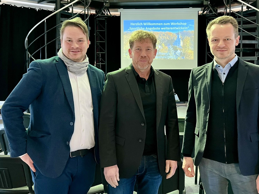 KI generiert: Das Bild zeigt eine Präsentation oder ein Meeting in einem Raum mit mehreren Personen. Zwei Männer stehen vorne und sprechen zu einer Gruppe von Zuhörern, während auf einer Leinwand eine PowerPoint-Präsentation mit dem Titel "Kupferhalte Tausug" und dem Datum "26.03.2025" projiziert wird.