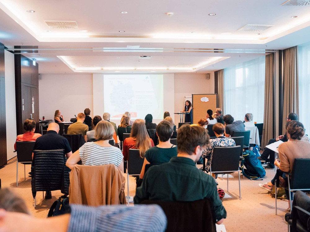 KI generiert: Das Bild zeigt eine Gruppe von Personen in einem Seminarraum, die einer Vortragenden zuhören, die an einem Pult steht. Der Raum ist hell und modern eingerichtet, mit großen Fenstern und einer Projektionsfläche im Hintergrund.