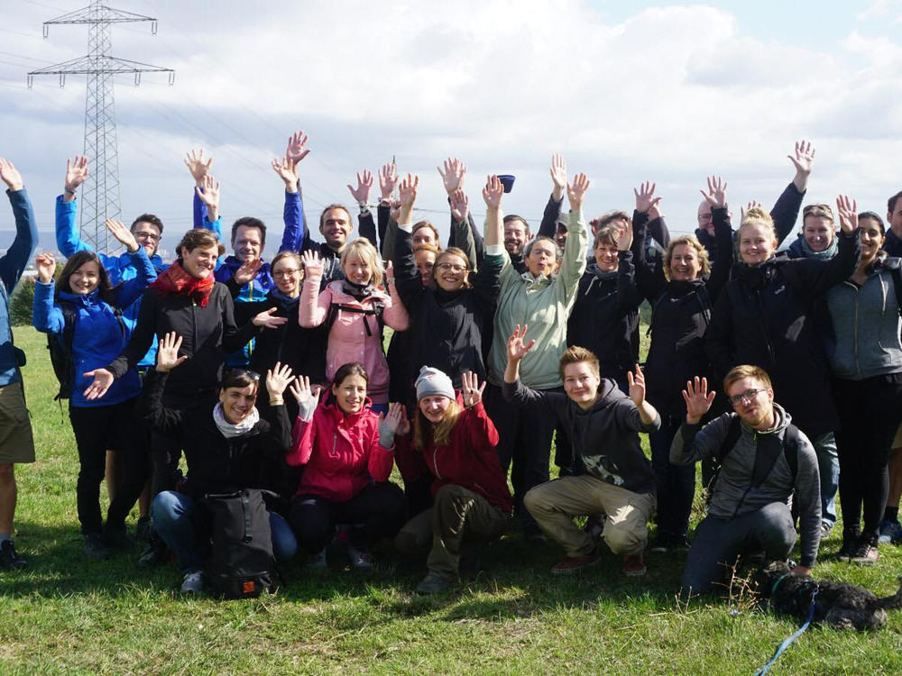 KI generiert: Das Bild zeigt eine Gruppe von Menschen, die draußen auf einer Wiese stehen und sich freudig mit hochgereckten Armen präsentieren. Im Vordergrund ist ein Hund zu sehen, und im Hintergrund sind Strommasten und eine leicht bewölkte Himmel zu erkennen.