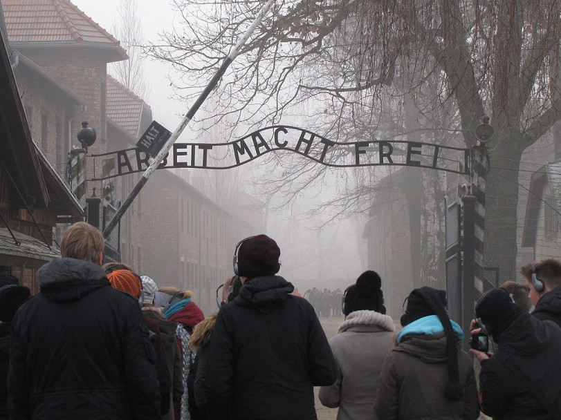 KI generiert: Das Bild zeigt eine Gruppe von Menschen, die durch das Tor mit der Aufschrift "Arbeit Macht Frei" am Eingang des ehemaligen Konzentrationslagers Auschwitz geht. Der Hauptinhalt des Bildes ist die symbolträchtige und historische Bedeutung des Ortes, der an die Gräueltaten des Holocaust erinnert.