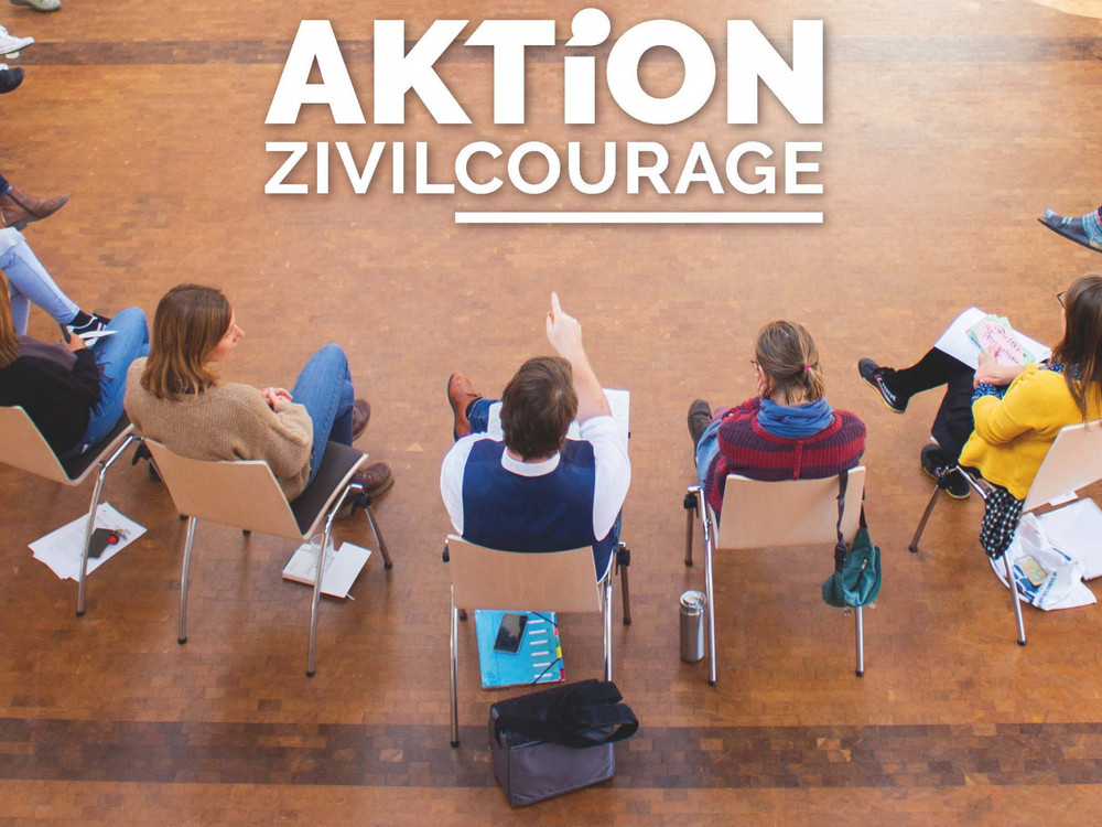 KI generiert: Das Bild zeigt eine Gruppe von Menschen, die in einem Kreis auf dem Boden sitzen und sich anscheinend unterhalten oder einer Aktivität widmen. Im Zentrum des Bildes steht der Text "AKTiON ZIVILCOURAGE".