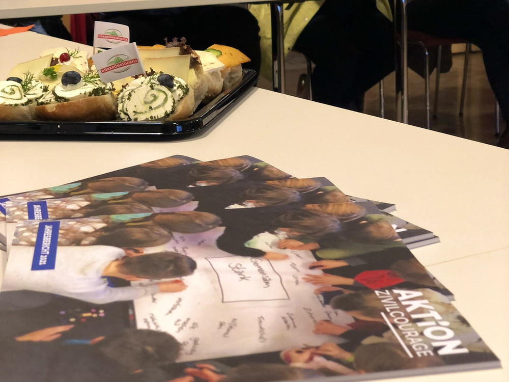 KI generiert: Das Bild zeigt eine Gruppe von Personen, die an langen Tischen in einem Raum mit großen Fenstern sitzen und sich in einer Besprechung oder einem Meeting befinden. Der Hauptinhalt des Bildes ist eine formelle Zusammenkunft oder Konferenz.