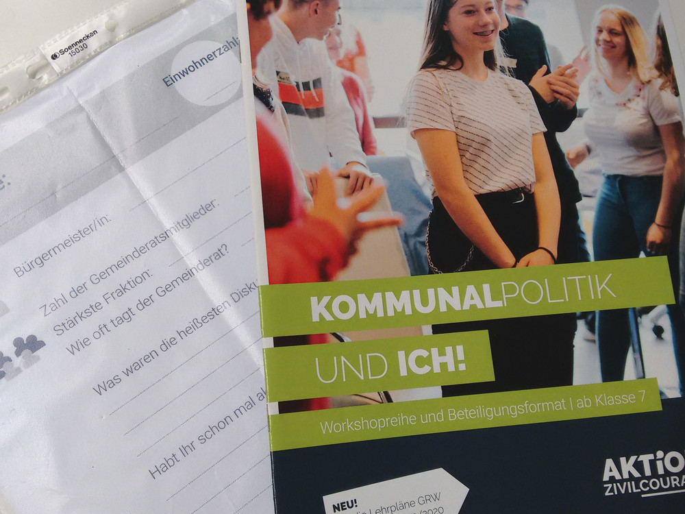 KI generiert: Das Bild zeigt ein Heft mit dem Titel "Kommunalpolitik und ich!" auf der Titelseite, das sich an Schüler ab Klasse 7 richtet und von der Aktion Zivilcourage herausgegeben wird. Neben dem Heft liegt ein leeres Blatt Papier mit einigen handschriftlichen Notizen und einem Stempel.
