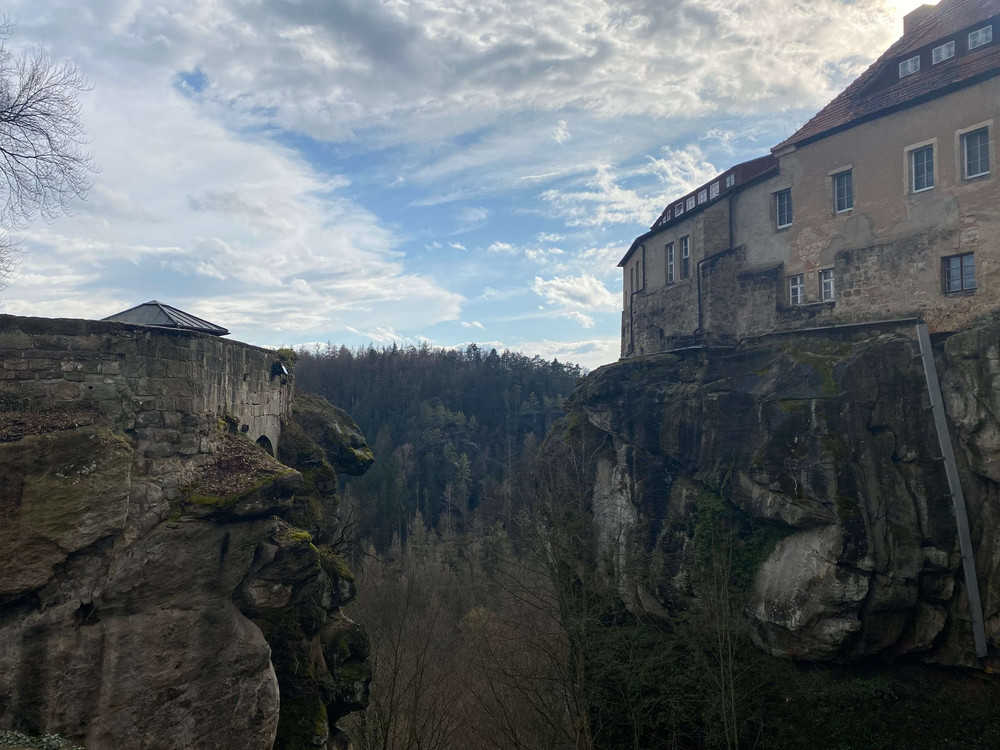 KI generiert: Das Bild zeigt eine alte Burg oder Festung, die auf einem Felsen am Rand eines tiefen Waldtals thront. Der Hauptinhalt des Bildes ist die beeindruckende Architektur der Burg und die malerische Landschaft mit dem Wald und dem Himmel im Hintergrund.