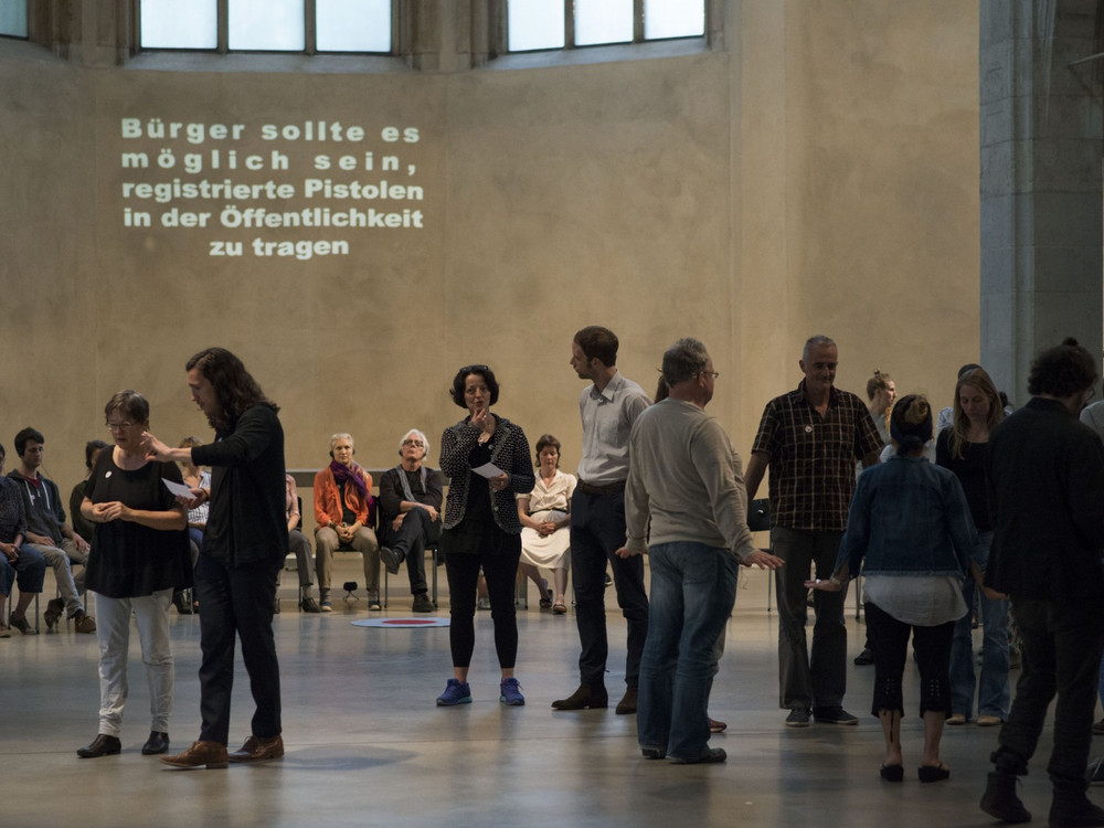 KI generiert: Das Bild zeigt eine Gruppe von Menschen in einer großen, hallenartigen Umgebung, die sich zu unterhalten und zu diskutieren scheinen. Im Hintergrund ist eine Projektion mit einem deutschen Text, der zur Registrierung von Pistolen in der Öffentlichkeit auffordert.