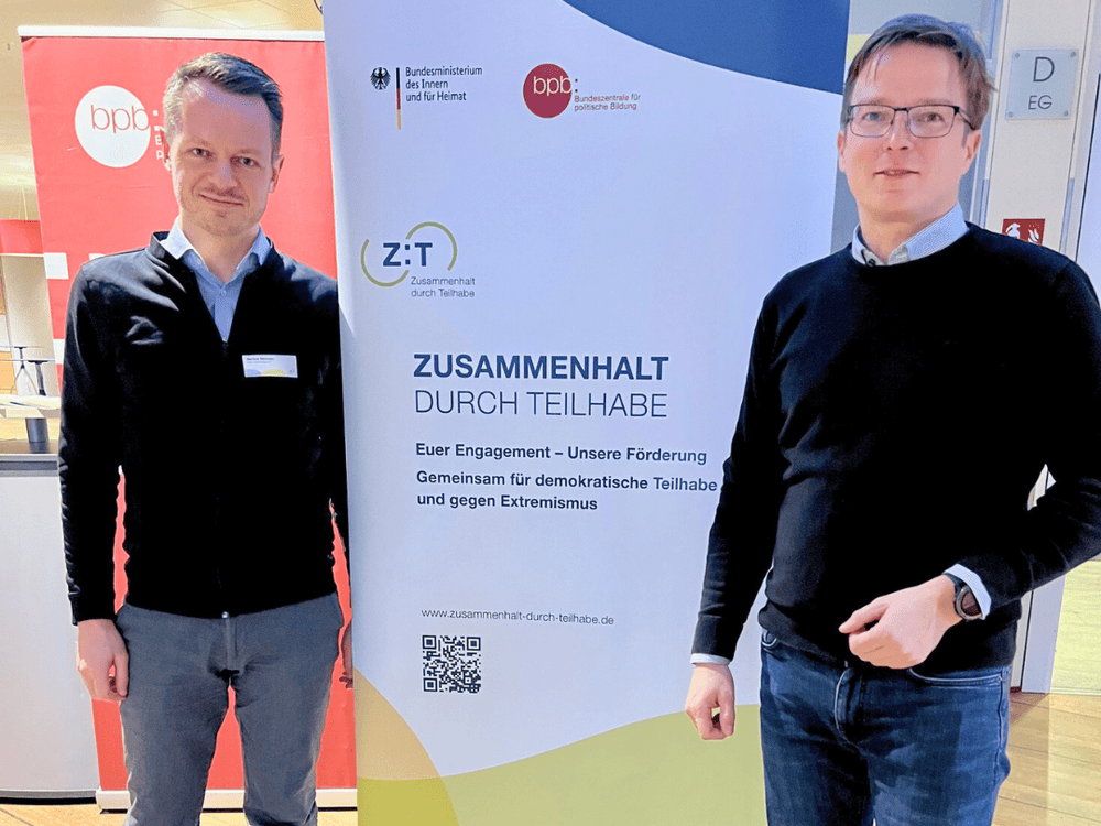 KI generiert: Das Bild zeigt eine Vortragsveranstaltung in einem großen, gut besuchten Hörsaal. Eine Person hält einen Vortrag auf der Bühne, während das Publikum in blauen Sitzen zuhört und zwei Projektionsleinwände mit Präsentationen im Hintergrund sichtbar sind.